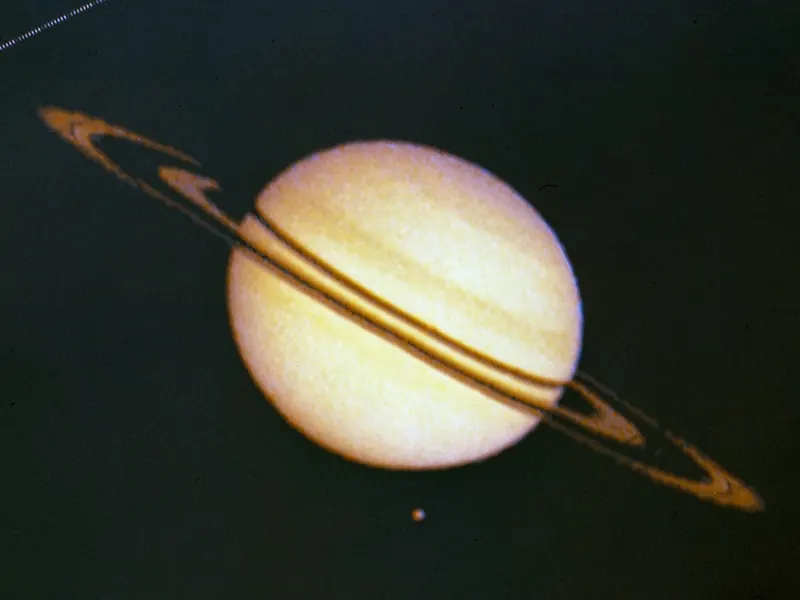 A grainy, blue-tinted picture of Saturn and its rings with a small moon beside it
